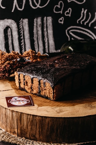 A rich chocolate cake sits on a wooden slab, surrounded by rustic decorations and chalkboard-style illustrations of food and hearts in the background. A label reading 'Delicias da Nila' is displayed in the foreground.