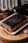 A rectangular chocolate cake is presented on a white plate. The cake is heavily coated with dark chocolate icing that drips down the sides. It sits on a wooden surface, accompanied by a dried floral arrangement and a wine bottle lying horizontally. A business card with a logo and text is placed next to the cake.
