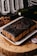 A rectangular chocolate cake is presented on a white plate. The cake is heavily coated with dark chocolate icing that drips down the sides. It sits on a wooden surface, accompanied by a dried floral arrangement and a wine bottle lying horizontally. A business card with a logo and text is placed next to the cake.