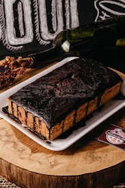 A rectangular chocolate cake is presented on a white plate. The cake is heavily coated with dark chocolate icing that drips down the sides. It sits on a wooden surface, accompanied by a dried floral arrangement and a wine bottle lying horizontally. A business card with a logo and text is placed next to the cake.