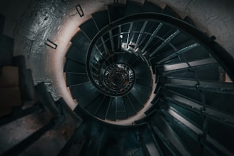 A spiral staircase viewed from above, showcasing a series of dark steps winding downward, with metal railings curving along the contour. The central axis of the staircase creates a hypnotic, tunnel-like appearance.