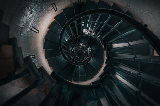 A spiral staircase viewed from above, showcasing a series of dark steps winding downward, with metal railings curving along the contour. The central axis of the staircase creates a hypnotic, tunnel-like appearance.