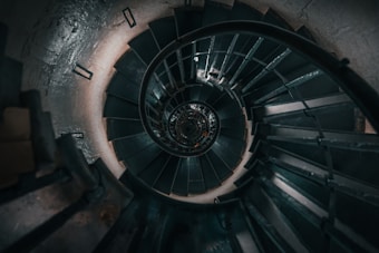 A spiral staircase viewed from above, showcasing a series of dark steps winding downward, with metal railings curving along the contour. The central axis of the staircase creates a hypnotic, tunnel-like appearance.
