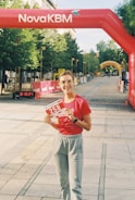 A person is standing on a pedestrian pathway holding a race bib numbered 258. They are wearing a red shirt and gray sweatpants. Above them, there's an inflatable red arch with the text NovaKBM. The pathway is lined with trees and barriers, indicating a race event. Banners and another yellow arch can be seen in the distance.