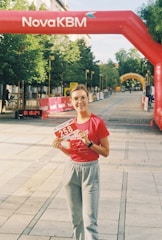 A person is standing on a pedestrian pathway holding a race bib numbered 258. They are wearing a red shirt and gray sweatpants. Above them, there's an inflatable red arch with the text NovaKBM. The pathway is lined with trees and barriers, indicating a race event. Banners and another yellow arch can be seen in the distance.