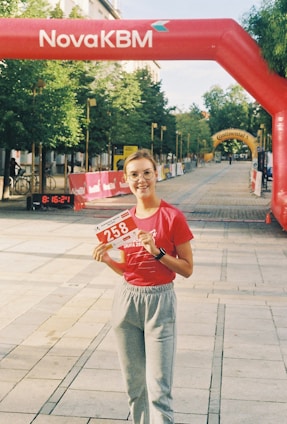 A person is standing on a pedestrian pathway holding a race bib numbered 258. They are wearing a red shirt and gray sweatpants. Above them, there's an inflatable red arch with the text NovaKBM. The pathway is lined with trees and barriers, indicating a race event. Banners and another yellow arch can be seen in the distance.
