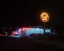 A nighttime street view featuring a brightly lit neon sign for 'Peppermint Hippo' in the foreground. In the background, there is a building with illuminated red signage for 'Boston Pizza'. Several parked cars are lined up along the side of the building, with minimal street lighting enhancing the nighttime ambiance.