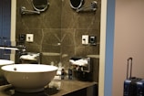 A modern bathroom setup with a white basin on a marble countertop. There is a chrome faucet next to the sink. Above the sink, a mirrored wall reflects the light, and there is a mounted magnifying mirror. To the side, there is a tissue dispenser and toiletries organized on the countertop. A blue suitcase stands in the corner next to the wall.