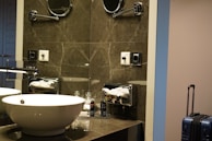 A modern bathroom setup with a white basin on a marble countertop. There is a chrome faucet next to the sink. Above the sink, a mirrored wall reflects the light, and there is a mounted magnifying mirror. To the side, there is a tissue dispenser and toiletries organized on the countertop. A blue suitcase stands in the corner next to the wall.