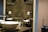 A modern bathroom setup with a white basin on a marble countertop. There is a chrome faucet next to the sink. Above the sink, a mirrored wall reflects the light, and there is a mounted magnifying mirror. To the side, there is a tissue dispenser and toiletries organized on the countertop. A blue suitcase stands in the corner next to the wall.