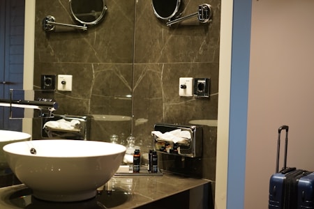 A modern bathroom setup with a white basin on a marble countertop. There is a chrome faucet next to the sink. Above the sink, a mirrored wall reflects the light, and there is a mounted magnifying mirror. To the side, there is a tissue dispenser and toiletries organized on the countertop. A blue suitcase stands in the corner next to the wall.