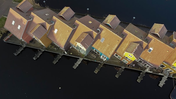 Aerial view of colorful, connected houses with gabled roofs, situated on the edge of a dark body of water. The houses have varied shades of yellow, orange, and red, creating a lively pattern. Wooden docks are positioned in front of each house, stretching out into the water. The surrounding area appears serene and orderly.