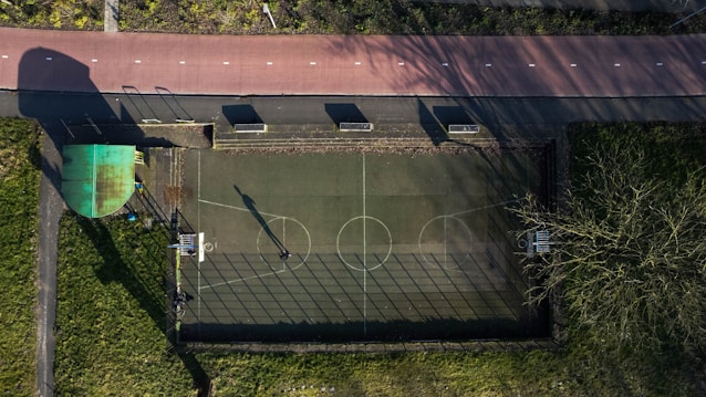 An aerial view of a small outdoor basketball court with clear markings. The court is fenced and located next to a paved path with bike lanes. Shadows are cast over the area, indicating a low sun angle. Surrounding areas include grass, trees, and some pavement.