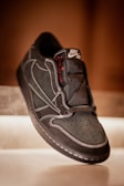 A close-up view of a black sneaker featuring white stitching and a visible Nike Air logo on the tongue. The sneaker has a perforated toebox and a text label with the word 'Cactus' in red. The shoe is positioned against a warm, softly lit background, giving it a luxurious appearance.