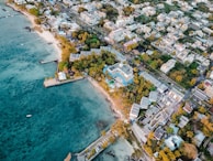 An aerial view of a coastal town featuring a mix of residential and commercial buildings surrounded by lush greenery. The coastline is lined with sandy beaches and turquoise waters, with a few docks extending into the ocean. The area is populated with palm trees, adding a tropical feel to the scene.