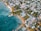 An aerial view of a coastal town featuring a mix of residential and commercial buildings surrounded by lush greenery. The coastline is lined with sandy beaches and turquoise waters, with a few docks extending into the ocean. The area is populated with palm trees, adding a tropical feel to the scene.
