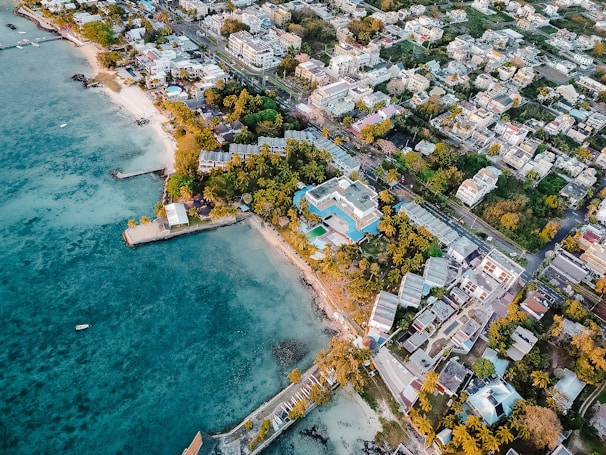 An aerial view of a coastal town featuring a mix of residential and commercial buildings surrounded by lush greenery. The coastline is lined with sandy beaches and turquoise waters, with a few docks extending into the ocean. The area is populated with palm trees, adding a tropical feel to the scene.