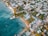 An aerial view of a coastal town featuring a mix of residential and commercial buildings surrounded by lush greenery. The coastline is lined with sandy beaches and turquoise waters, with a few docks extending into the ocean. The area is populated with palm trees, adding a tropical feel to the scene.