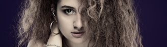 A person with curly brown hair partially covering one eye, wearing hoop earrings and a bracelet, against a dark purple background.