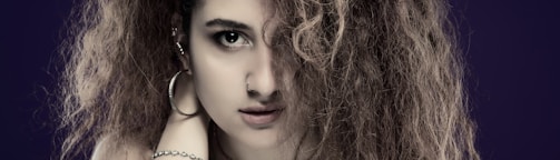 A person with curly brown hair partially covering one eye, wearing hoop earrings and a bracelet, against a dark purple background.