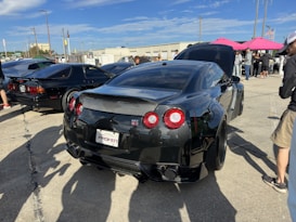 A black sports car with a sleek design is parked in an outdoor gathering. Several people are standing around, some of them inspecting the vehicles. The car's license plate reads 'PROVEN' and another black car is visible to the left. The sky is clear with a few clouds and the concrete ground reflects the sunlight.