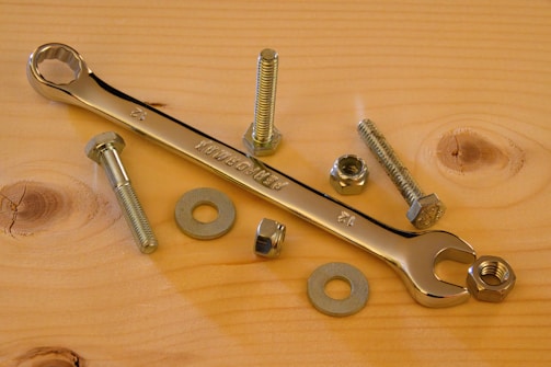 A flat wooden surface hosts a collection of metal hardware items, including a shiny chrome wrench, several bolts, nuts, and washers. The arrangement exhibits an orderly yet casual layout, with natural wood grain patterns visible in the background.