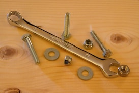 A flat wooden surface hosts a collection of metal hardware items, including a shiny chrome wrench, several bolts, nuts, and washers. The arrangement exhibits an orderly yet casual layout, with natural wood grain patterns visible in the background.