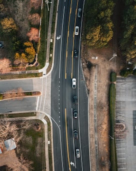 An aerial view of a road with multiple cars driving in a line. The road curves slightly to the right and is surrounded by various landscapes including grassy areas, trees with autumn foliage, and paved surfaces such as driveways and parking lots.