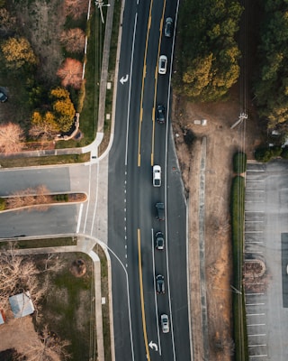 An aerial view of a road with multiple cars driving in a line. The road curves slightly to the right and is surrounded by various landscapes including grassy areas, trees with autumn foliage, and paved surfaces such as driveways and parking lots.