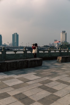 A person wearing a white jacket with a red hood and holding a camera stands on a stone platform near a waterfront. The background showcases a cityscape with tall buildings and a bridge. The foreground is a tiled walkway with a cloudy sky above.