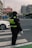 A traffic officer wearing a dark uniform with reflective stripes stands at a crosswalk holding a white baton. The setting is an urban street with a white car nearby and city buildings in the background. There are signs in Chinese characters and the sky appears overcast.