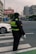 A traffic officer wearing a dark uniform with reflective stripes stands at a crosswalk holding a white baton. The setting is an urban street with a white car nearby and city buildings in the background. There are signs in Chinese characters and the sky appears overcast.