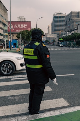 A traffic officer wearing a dark uniform with reflective stripes stands at a crosswalk holding a white baton. The setting is an urban street with a white car nearby and city buildings in the background. There are signs in Chinese characters and the sky appears overcast.