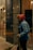 A person dressed in warm clothing, including a red hooded jacket with fur trim and a blue coat, holding a broom. The individual appears to be cleaning outside a building with a glass facade at night.
