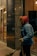 A person dressed in warm clothing, including a red hooded jacket with fur trim and a blue coat, holding a broom. The individual appears to be cleaning outside a building with a glass facade at night.