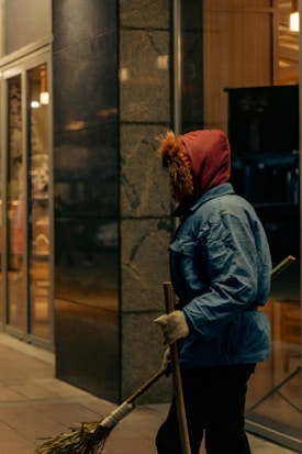 A person dressed in warm clothing, including a red hooded jacket with fur trim and a blue coat, holding a broom. The individual appears to be cleaning outside a building with a glass facade at night.