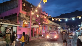 A vibrant street scene during dusk in a small town with colorful buildings. The prominent pink building on the left has a rustic, artistic design and a sign that reads 'Temazcal'. Strings of decorative lights hang across the street, illuminating the cobblestone road. Several people stroll down the street, and a car is parked in the center. The mountainous backdrop adds to the scenic atmosphere of the area.
