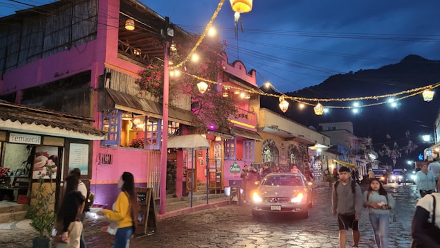 A vibrant street scene during dusk in a small town with colorful buildings. The prominent pink building on the left has a rustic, artistic design and a sign that reads 'Temazcal'. Strings of decorative lights hang across the street, illuminating the cobblestone road. Several people stroll down the street, and a car is parked in the center. The mountainous backdrop adds to the scenic atmosphere of the area.