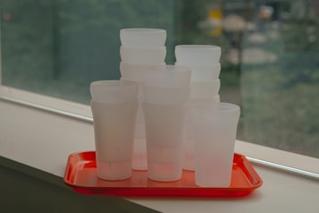 A stack of frosted plastic cups is neatly arranged on a bright red tray, placed on a windowsill. The background is a soft focus of greenery, seen through the window, providing a contrast to the sharp focus of the cups and tray.