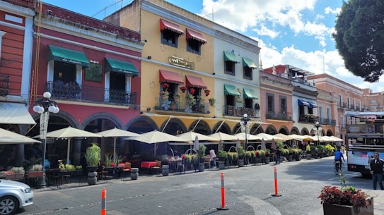 A charming streetscape featuring colorful, historic buildings with arched doorways and balconies adorned with plants. Caf&eacute; or restaurant patios line the sidewalk, shaded by large umbrellas. People walk and cycle along the street, while a tree provides greenery against the backdrop of a vibrant, sunny sky.