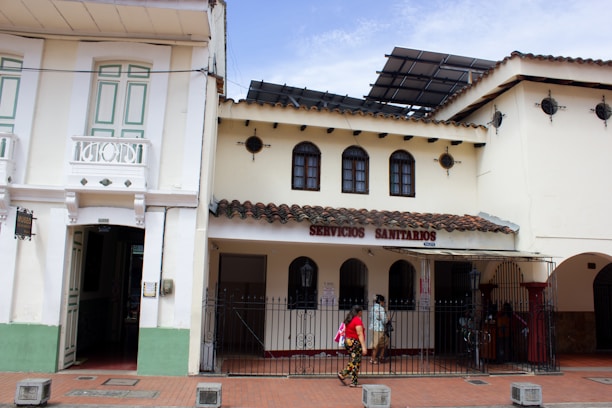 A street view featuring a traditional building with white walls and a tiled roof. The structure on the left has detailed woodwork and pastel green shutters, while the building on the right displays a sign reading 'SERVICIOS SANITARIOS'. A few people are walking past under a partially cloudy sky.