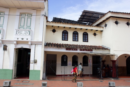 A street view featuring a traditional building with white walls and a tiled roof. The structure on the left has detailed woodwork and pastel green shutters, while the building on the right displays a sign reading 'SERVICIOS SANITARIOS'. A few people are walking past under a partially cloudy sky.