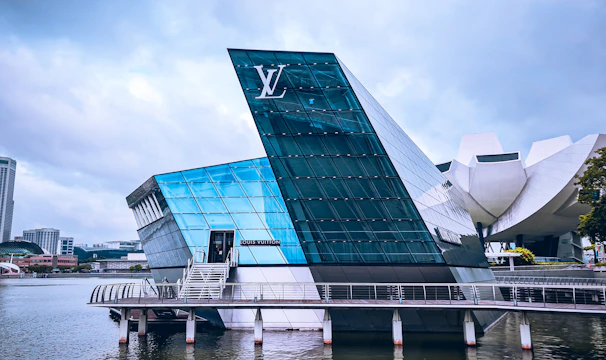 A modern glass building with angular architecture is situated on the water. The structure has a prominent logo and features reflective blue and green glass surfaces. Nearby, a curved white building is partially visible, and there are cityscape elements in the background under a cloudy sky.