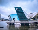 A modern glass building with angular architecture is situated on the water. The structure has a prominent logo and features reflective blue and green glass surfaces. Nearby, a curved white building is partially visible, and there are cityscape elements in the background under a cloudy sky.