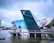 A modern glass building with angular architecture is situated on the water. The structure has a prominent logo and features reflective blue and green glass surfaces. Nearby, a curved white building is partially visible, and there are cityscape elements in the background under a cloudy sky.