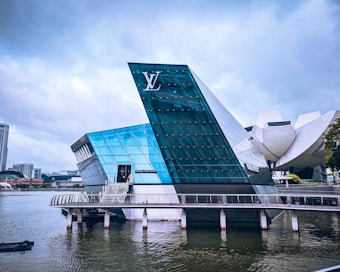 A modern glass building with angular architecture is situated on the water. The structure has a prominent logo and features reflective blue and green glass surfaces. Nearby, a curved white building is partially visible, and there are cityscape elements in the background under a cloudy sky.