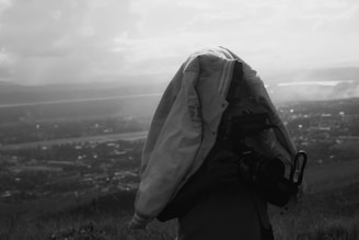A person is standing outdoors with a large camera covered by a jacket, obscuring their face. The background features a hazy, expansive landscape with hills and a body of water in the distance. It appears to be a grayscale or black-and-white photo.