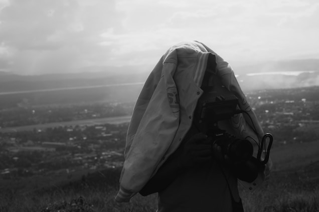 A person is standing outdoors with a large camera covered by a jacket, obscuring their face. The background features a hazy, expansive landscape with hills and a body of water in the distance. It appears to be a grayscale or black-and-white photo.