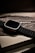 A sleek, modern smartwatch with a black ribbed band is placed on top of its packaging box, which shows an artistic wave design and the logo of the brand. The watch has a metallic casing and is presented at a slight angle, creating a sophisticated and luxurious appearance.