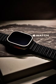 A sleek, modern smartwatch with a black ribbed band is placed on top of its packaging box, which shows an artistic wave design and the logo of the brand. The watch has a metallic casing and is presented at a slight angle, creating a sophisticated and luxurious appearance.